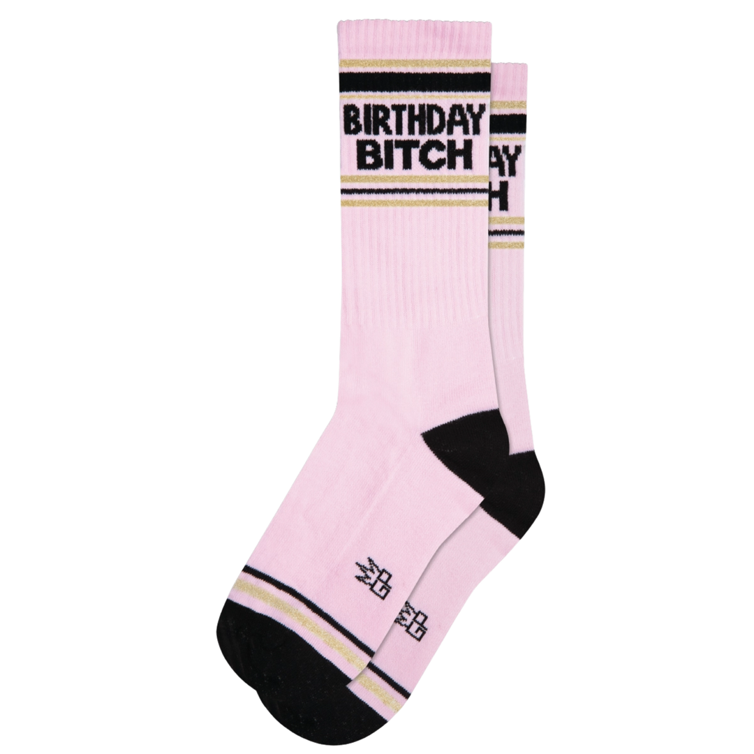 Pink socks with black and yellow stripes and black text reading "Birthday Bitch". When celebrating a special day, these socks offer plush comfort to the honored Birthday Bitch. Crafted from a blend of cotton, nylon, and spandex, they are proudly made in the USA and provide a unisex, one size fits most fit.